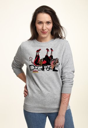 Kvinde med brunt hår bærer en grå sweatshirt med en tegneserie-Deadpool-design og blå jeans, står med den ene hånd på hoften.