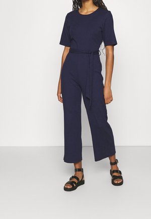 Marineblauwe geribbelde jumpsuit met korte mouwen, ronde hals en een taille met ceintuur. Wijde broekspijpen, gedragen met zwarte sandalen met bandjes.