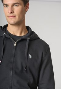 Felpa nera con zip realizzata in cotone, caratterizzata da un emblema argento sul lato sinistro, lacci a contrasto e chiusura zip frontale.