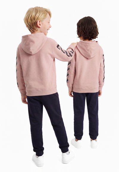 Две деца, облечени в розови суичъри с качулки и тъмни панталони, стоят с гръб една към друга; едното се усмихва и поставя ръка на рамото на другото.