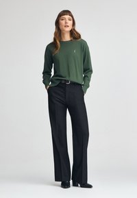 Haut vert à manches longues avec un col rond, associé à un pantalon noir taille haute et des bottes noires à talons. Détail du logo simple sur le haut.