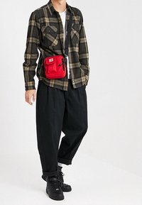 Plaid flannel shirt in black and olive, top pocket detailing, paired with loose black trousers and a red crossbody bag. Black sneakers worn.
