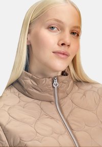 Beżowa pikowana kurtka z wysokim kołnierzem, srebrnym suwakem z uchwytem w kształcie serca oraz wzorem serc na powierzchni tkaniny.