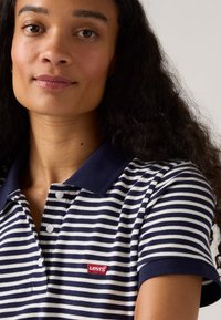 Mörkblå och vit randig polo shirt med mörkblå krage, prydd med en röd Levi's logotyppatch på vänster sida. Korta ärmar, knäppning fram.