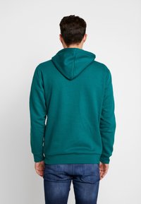Man med teal hoodie och blå jeans, stående vänd bort från en enfärgad vit bakgrund.