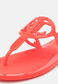 Korallfärgade gummiskor med texturerad fotbädd och en stor, iögonfallande logotyp på remmen. Designen har en sula i tångstil.