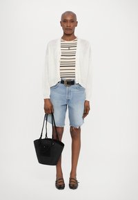 Weiße, offen gestrickte Cardigan über einem beige- und schwarzgestreiften Shirt, kombiniert mit hellblauen Denim-Shorts und schwarzen Ballerinas. Hält eine schwarze geflochtene Handtasche.