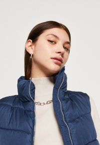 Ναυτικό επενδυμένο γιλέκο με ψηλό γιακά και ασημένιο φερμουάρ. Φοριέται πάνω από έναν μπεζ ψηλό λαιμό, με ασημένια αλυσίδα γύρω από τον λαιμό.