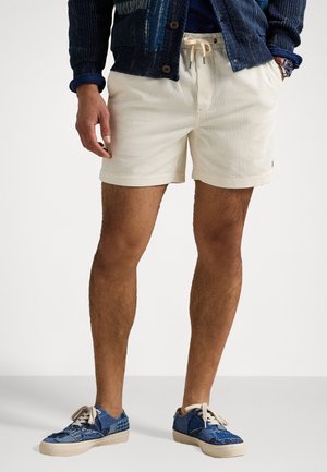 Homme portant un short crème à cordon de serrage, des baskets patchwork bleu marine et une veste texturée bleu foncé avec les mains dans les poches.