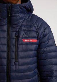 Marineblå puffjakke med quiltet tekstur, fuld lynlås, sorte lynlåse og en rød "JACK1T" logo accent på brystet.