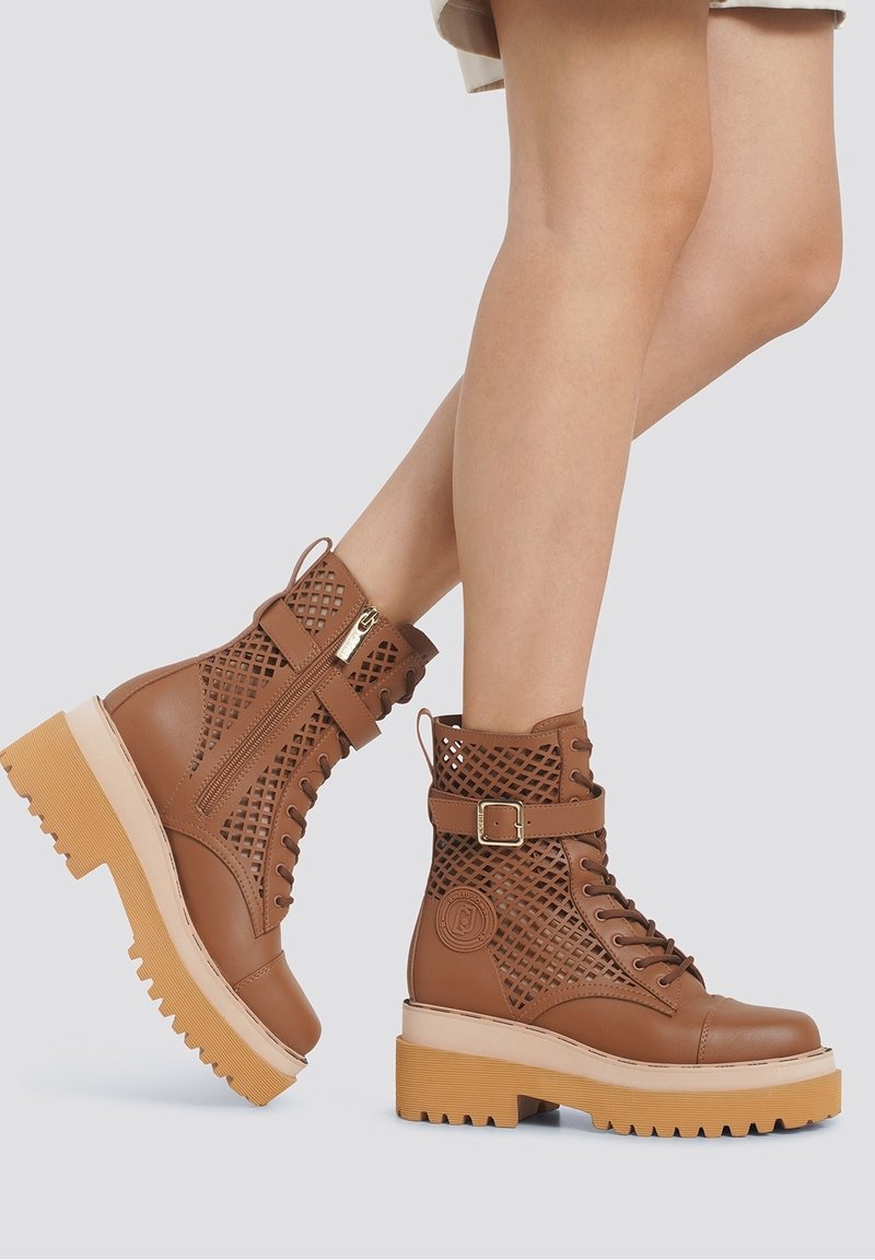 Bottes à lacets marron à la cheville avec boucle et fermeture éclair latérale, avec un motif en treillis et une semelle épaisse marron clair, portées sur des jambes croisées.