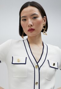 Bílý kardigan s krátkým rukávem a námořnickým lemem, dvěma předními kapsami, zlatými knoflíky a texturovanou tkaninou. Modelka nosí zlaté geometrické náušnice.