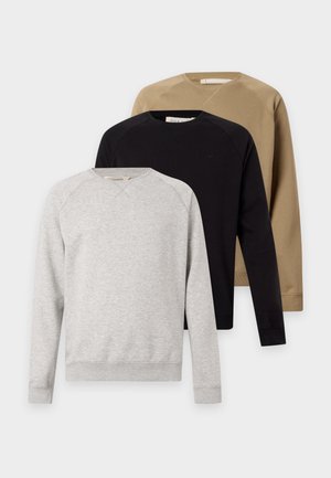 Je jeune homme debout, portant une casquette grise, un sweat-shirt noir, un pantalon blanc et des baskets noires sur un fond blanc uni.
