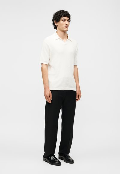 Άνδρας όρθιος, φοράει κοντομάνικη λευκή πλεκτή πόλο, μαύρο παντελόνι και μαύρα δερμάτινα μοκασίνια σε απλό ανοιχτό φόντο.