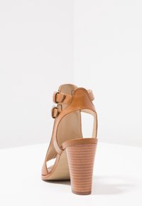 Korkean korotuksen tan-nahkainen sandaali, jossa on kaareva leikkaussuunnittelu, säädettävät soljet ja teksturoitu puukorko.