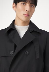 Čierny trench coat s dvojradovými gombíkmi, širokým golierom a hladkou, jemnou textúrou. Nosený cez svetlý pletený sveter.