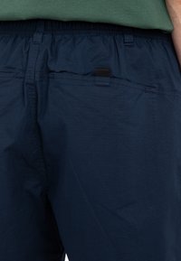 Navy blue shorts made from lightweight fabric, featuring an elastic waistband, side pocket, and a small black tab detail.