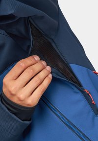 Niebieska kurtka z kieszenią na zamek, prezentująca teksturowany, oddychający materiał. Kolory akcentowe to granatowe i pomarańczowe detale. Ręka sięga do kieszeni.