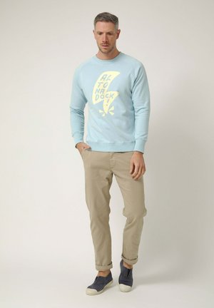 Hombre con sudadera azul claro con el texto "Altona Dock", pantalones beige y zapatos sin cordones azul marino, de pie contra un fondo liso.