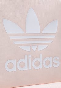Růžová tkaninová taška s velkým bílým logem Adidas, které obsahuje tři lístkovité prvky a vodorovné pruhy, texturovaný materiál.