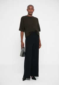 Donkergroen oversized gebreide top, brede zwarte broeken en puntige zwarte ballerina's. Model houdt een kleine zwarte handtas vast en staat tegen een effen achtergrond.