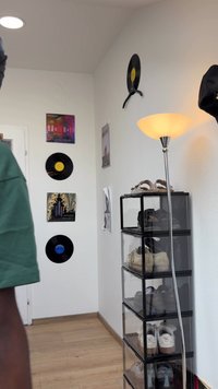 Organizador de sapatos em plástico transparente com cinco prateleiras, segurando vários calçados. A parede apresenta discos de vinil pretos e obras de arte coloridas; lâmpada de chão prateada incluída.