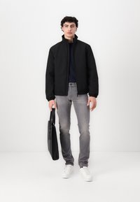 Sort, lynlåset jakke med høj krave, parret med en marineblå skjorte, lysegrå jeans og hvide sneakers. Holder en sort skuldertaske.