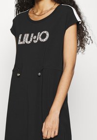 Czarna sukienka krótko rękawowa z okrągłym dekoltem, ozdobiona logo "LIU JO" z kryształów. Posiada sznurki w talii oraz kieszenie.