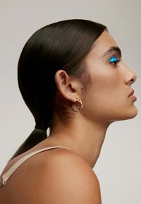 Pendientes de aro dorados con un diseño texturizado, llevados por una persona con cabello oscuro y liso y un maquillaje de ojos azul brillante, mostrada de perfil.