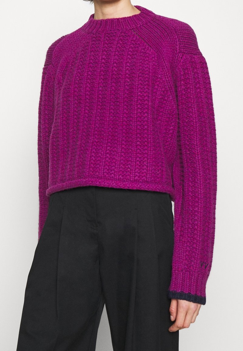 Person in einem strukturierten, magentafarbenen Strickpullover mit langen Ärmeln und schwarzen, weiten Hosen, von Hals bis Hüfte gezeigt.