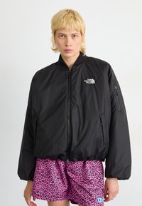 Zwarte bomberjack met een ritssluiting aan de voorkant, ribbelkraag en logo op de borst; gedragen over roze luipaardprint shorts. Soepele, lichte stof.