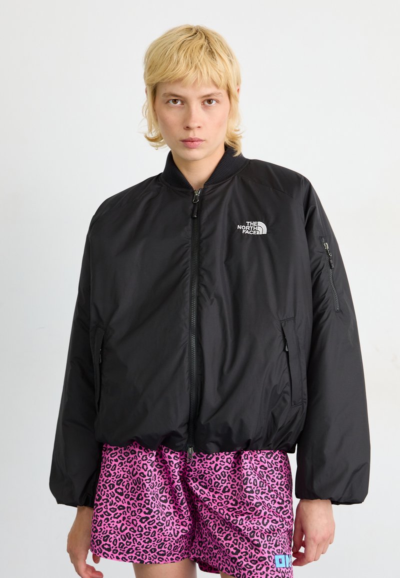 Zwarte bomberjack met een ritssluiting aan de voorkant, ribbelkraag en logo op de borst; gedragen over roze luipaardprint shorts. Soepele, lichte stof.