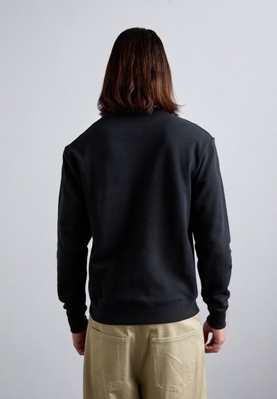 Black crew neck sweatshirt with long sleeves, textured fabric, and ribbed cuffs, paired with light beige trousers featuring back pockets.