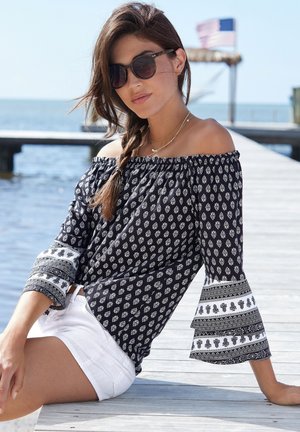 Schulterfreies Blusenshirt in Marineblau mit weißen Blumenmustern, mit Glockenärmel und Zierdetails; kombiniert mit weißen Shorts.