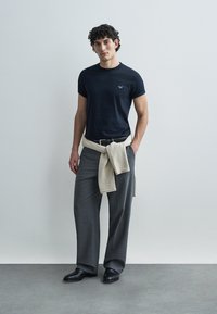 Marinblå t-shirt med rund hals, grå byxor med vida ben, beige tröja knuten runt midjan, svarta skor, slät tyg, minimal logodetalj på tröjan.