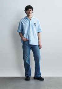 Jeune homme debout contre un mur uni, portant une chemise bleue claire à manches courtes avec un blason, un jean bleu et des chaussures noires.