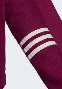 Ärm av ett vinrött plagg med tre parallella vita ränder, som visar detaljer av den texturerade tyget och sömmarna på en enfärgad bakgrund.