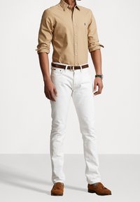 Camisa beige de botones con mangas remangadas, jeans blancos y mocasines marrones. Los acentos notables incluyen un pequeño logo en la camisa y un cinturón marrón.