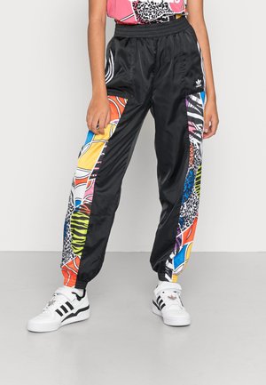 Pantalon de survêtement Adidas noir avec des panneaux latéraux à imprimé animal abstrait coloré, porté avec des baskets blanches Adidas et un haut rose à motifs.