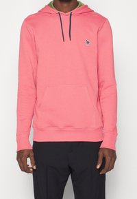 Homme portant un sweat à capuche rose vif avec poche avant et cordons de serrage bleu marine, associé à un pantalon noir, visible du cou jusqu'aux hanches.