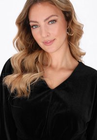 Fekete bársony ruhadarab mély V-nyakkivágással és bő, áramló ujjakkal. A szövet lágy tapintású, lazán hullámos haj egészíti ki.