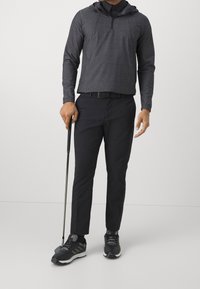 Pull-over noir à manches longues avec capuche, fermeture éclair sur le devant, associé à un pantalon noir et des chaussures de golf noires à motifs, tenant un club de golf.
