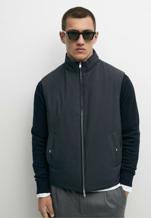 Uomo che indossa un gilet nero senza maniche con zip sopra una camicia nera a maniche lunghe, pantaloni grigi e occhiali da sole neri su sfondo semplice.