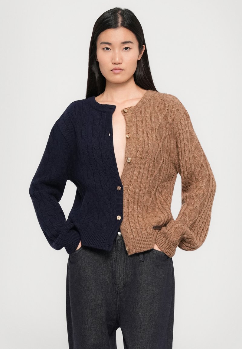 Cardigan bicolore con design a trecce. Lato sinistro blu navy con texture a coste, lato destro marrone chiaro con bottoni in legno, vestibilità rilassata.