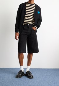 Černý cardigan s modrou výšivkou, pruhovaná béžová a černá košile, černé džínové šortky a černé boty s dekorativními akcenty.