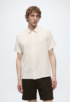 Ung mand med lyst bølget hår iført en tekstureret off-white kortærmet knappeskjorte og mørkebrune shorts, stående mod en ensfarvet baggrund.