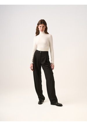 Bílý rolák s dlouhými rukávy v kombinaci s černými kalhotami s vysokým pasem a skladbami. Černé boty s texturovaným povrchem doplňují celkový outfit.
