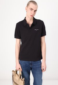 Polo negru cu mâneci scurte și guler. Prezintă un mic logo alb "TOMMY JEANS" pe partea stângă a pieptului. Este purtat cu blugi albaștri și ține o geantă bej.