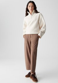 Weiße Fleece-Pullover mit hohem Kragen und halbem Reißverschluss, kombiniert mit braunen Tailored-Hosen und braunen Sneakers. Glatte Textur, lockere Passform.