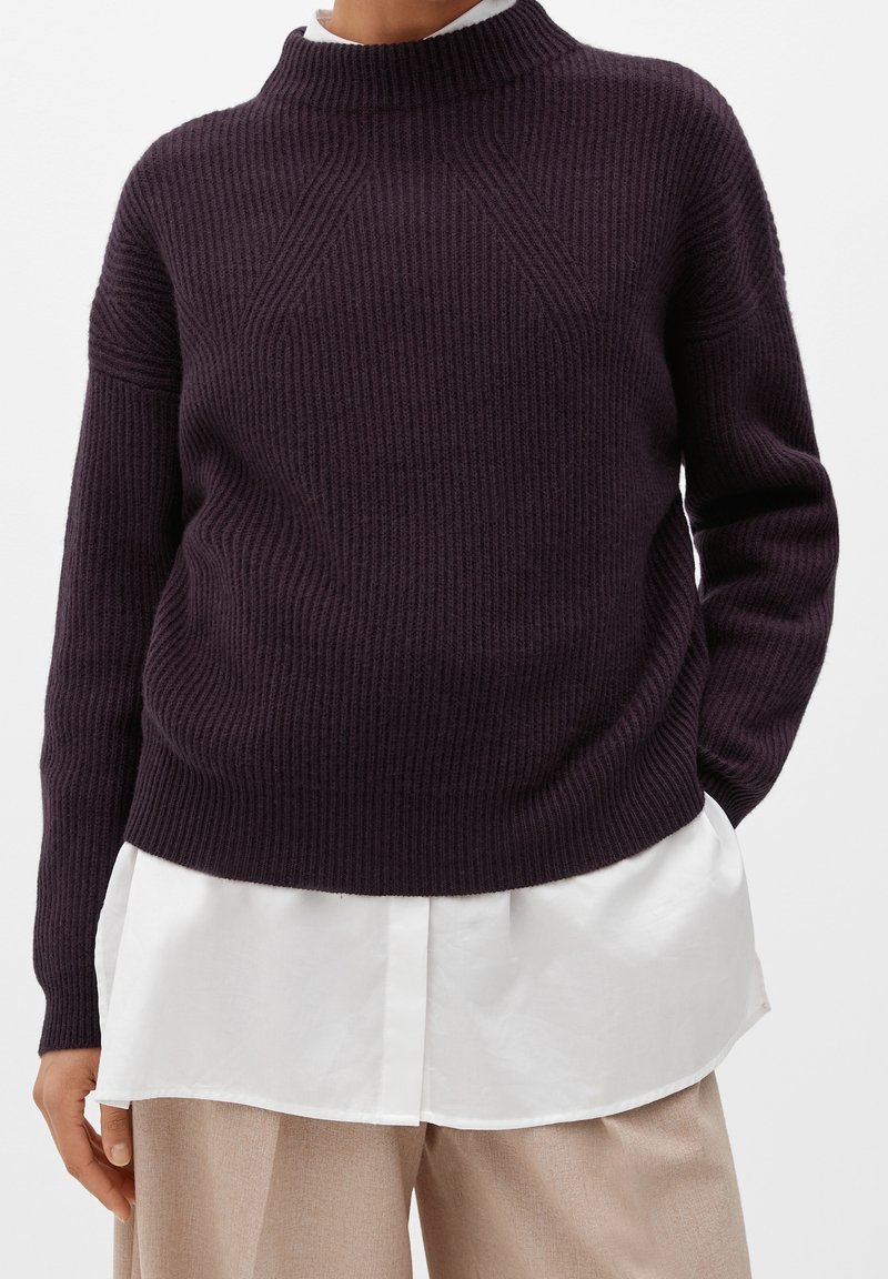 Pull côtelé violet foncé porté par-dessus une chemise blanche, associé à un pantalon beige, illustrant un style décontracté en couches.